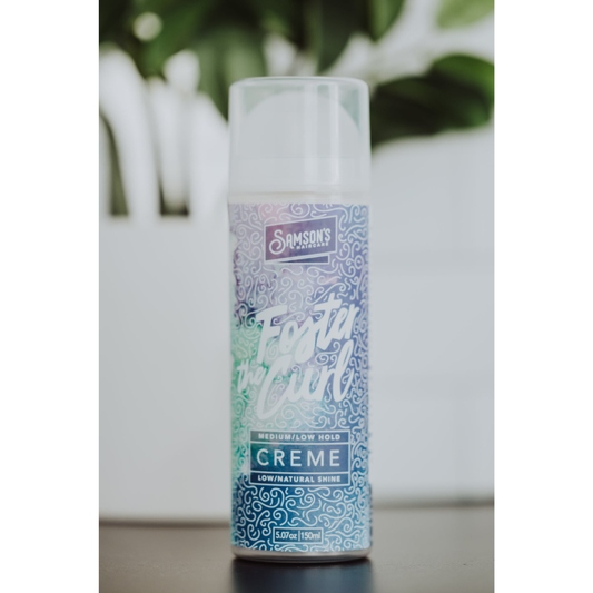 A 100ml can of Samson's Haircare Foster Curl Creme Medium/low hold hair spray, standing upright on a dark surface with a blurred white planter and green leaves in the background.