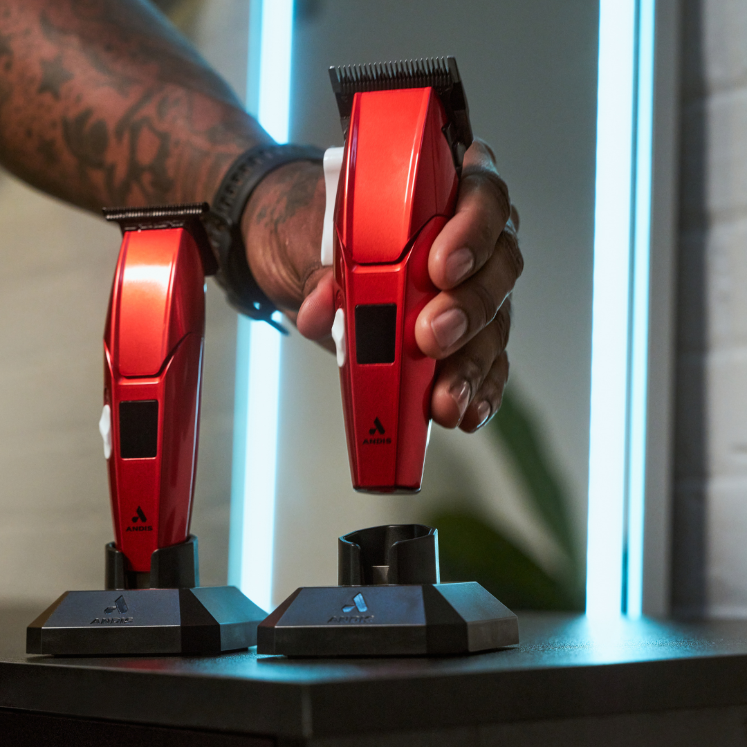 A tattooed hand holds a red and black clipper above two matching clipper stands on a dark tabletop, with a softly blurred indoor background.