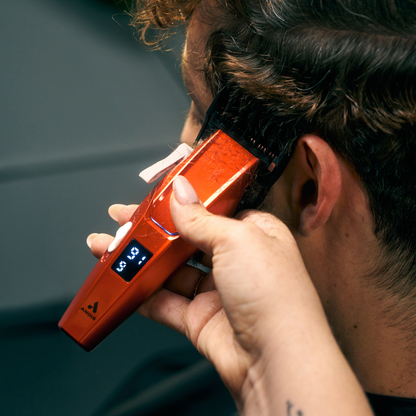 A close-up of a person using an orange electric hair clipper to trim their hair, with a small digital display on the clipper.